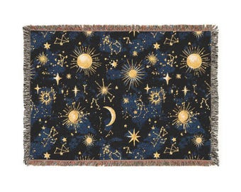 Peut inclure: Une couverture rectangulaire avec un motif céleste. Le fond est bleu foncé avec des soleils, des étoiles, des constellations et un croissant de lune dorés. Les bords sont frangés.