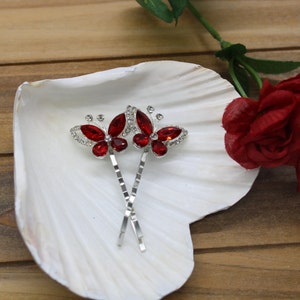 May include: Two silver butterfly hair clips with red rhinestone accents. The clips are on a white seashell-shaped surface.