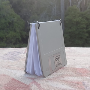 May include: A white 3.5 inch floppy disk with a white label that says "Verbatim Double Sided 3.5" and "HD" in black text. The disk is in a white cardboard sleeve with a black plastic binder clip.