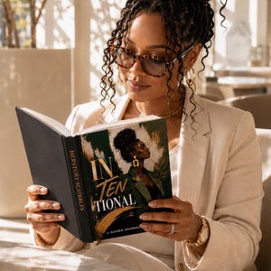 A woman with glasses and her hair in a high bun sits indoors while reading the open Intentional guided journal, wearing a light-colored blazer.