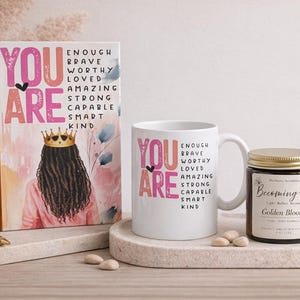 May include: A white ceramic mug, a notebook, and a candle in a brown glass jar with a gold lid. The mug and notebook feature the text YOU ARE ENOUGH BRAVE WORTHY LOVED AMAZING STRONG CAPABLE SMART KIND and an illustration of a person wearing a crown.