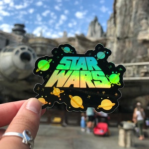 May include: A black holographic sticker with the text "STAR WARS" in a rainbow gradient. The sticker is surrounded by small yellow and green planets and stars.