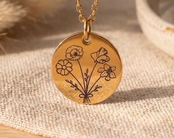 May include: A gold-toned metal necklace featuring a circular pendant with a delicate black line-art engraving of a small bouquet of wildflowers. The pendant hangs from a matching thin gold-toned chain, showcasing a polished, reflective surface.