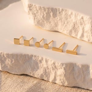 May include: A set of six gold-toned stud earrings featuring three distinct geometric shapes: two square studs, two circular studs, and two rectangular bar studs, all arranged in a row on a white textured surface.