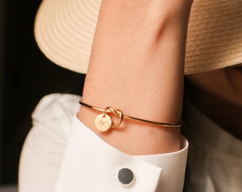 May include: A gold-colored bracelet with a knot design and a small circular charm. The charm features a hand gesture. The bracelet is worn on a wrist, paired with a white button-down shirt and a straw hat.