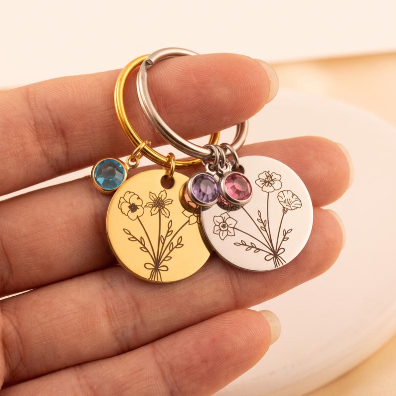 May include: Two circular metal keychains, one gold-toned and one silver-toned, each featuring an engraved floral bouquet design. The gold keychain includes a blue crystal charm, while the silver keychain features two small purple and pink crystal charms.