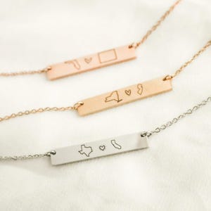 May include: Three gold bar necklaces with different state outlines and a heart charm. The necklaces are on a white surface.