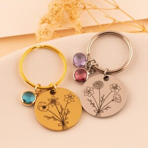 May include: Two circular metal keychains, one gold-toned and one silver-toned, each featuring a delicate line-art floral bouquet engraving. The gold keychain includes a single blue crystal charm, while the silver keychain features two crystal charms.
