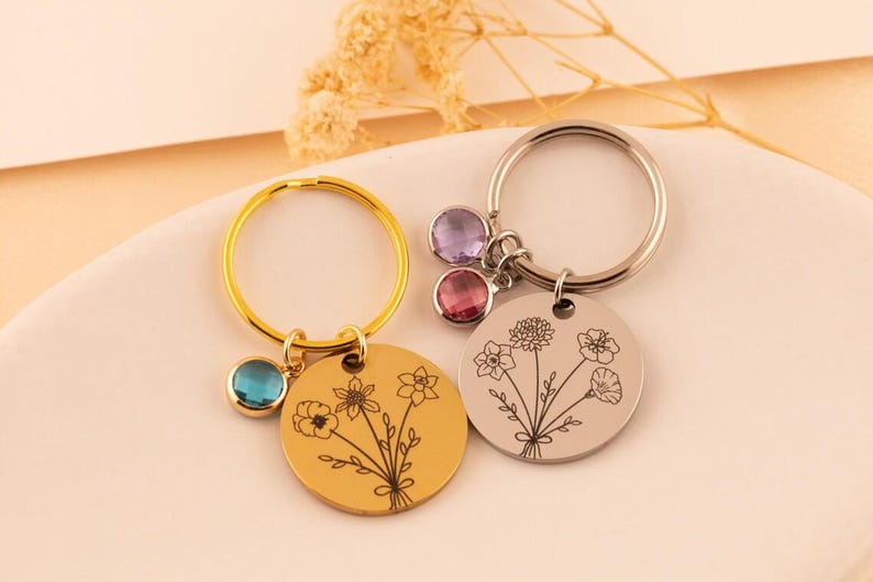 May include: Two circular metal keychains, one gold-toned and one silver-toned, each featuring a delicate line-art floral bouquet engraving. The gold keychain includes a single blue crystal charm, while the silver keychain features two crystal charms.