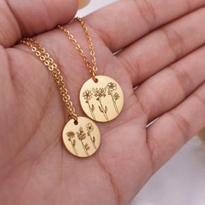 May include: Two gold-toned circular pendant necklaces featuring delicate black line art engravings of wildflowers. Each round charm hangs from a matching gold-toned chain, showcasing distinct botanical illustrations of various floral stems.