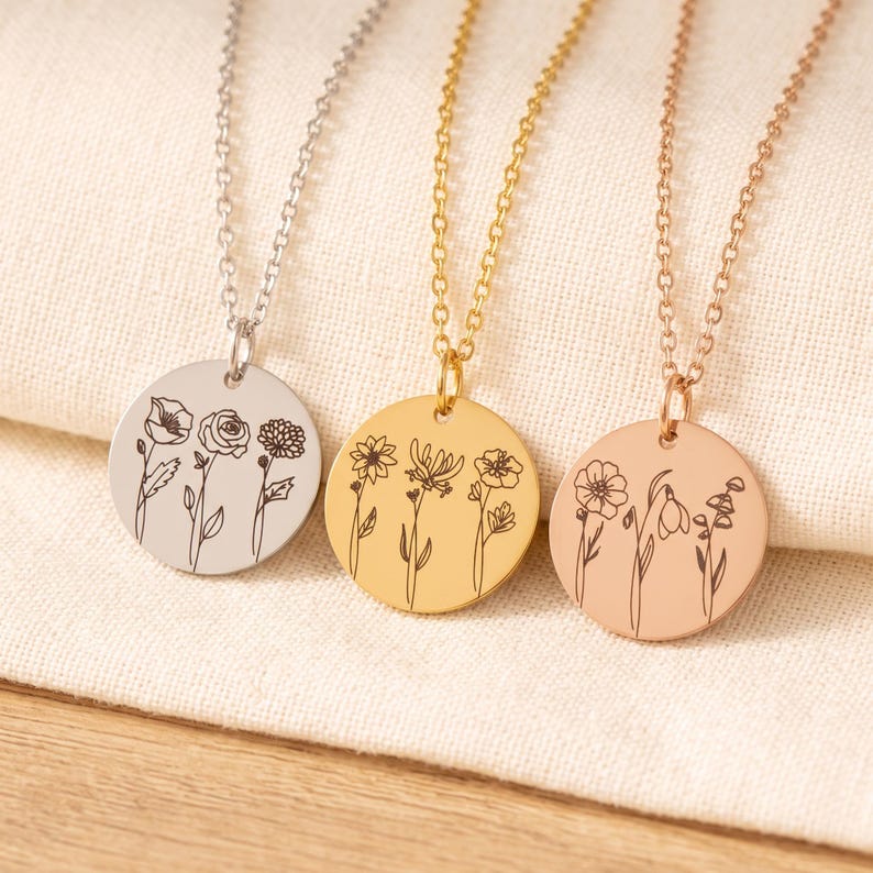 May include: Three circular pendant necklaces in silver, gold, and rose gold tones. Each pendant features a delicate black line art engraving of three distinct wildflowers. The necklaces are suspended from matching thin metal link chains.