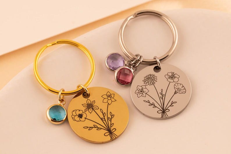 May include: Two circular metal keychains, one gold-toned and one silver-toned, each featuring a black line-art engraving of a flower bouquet. The gold keychain has a single blue crystal charm, while the silver keychain has two crystal charms.