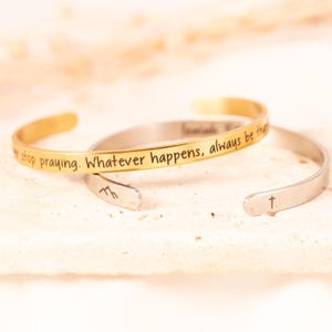 May include: Two open cuff bracelets. One is gold-toned with the inscription "...stop praying. Whatever happens, always be..." The other is silver-toned with a cross symbol. Both are on a light-colored surface.
