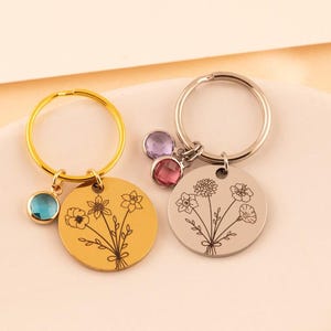 May include: Two circular metal keychains featuring engraved floral bouquet designs. One keychain is gold-toned with a single blue crystal charm, while the other is silver-toned with two crystal charms in purple and pink hues attached to the ring.