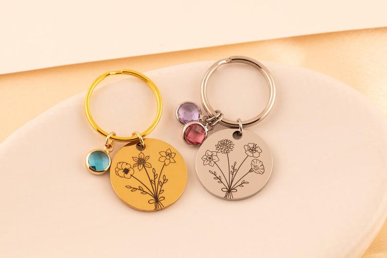 May include: Two circular metal keychains featuring engraved floral bouquet designs. One keychain is gold-toned with a single blue crystal charm, while the other is silver-toned with two crystal charms in purple and pink hues attached to the ring.