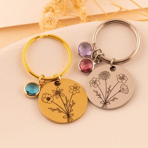 May include: Two circular metal keychains with engraved floral bouquets. One keychain is gold-toned with a blue crystal charm, and the other is silver-toned with two crystal charms in light purple and pink, featuring detailed line art of flowers.