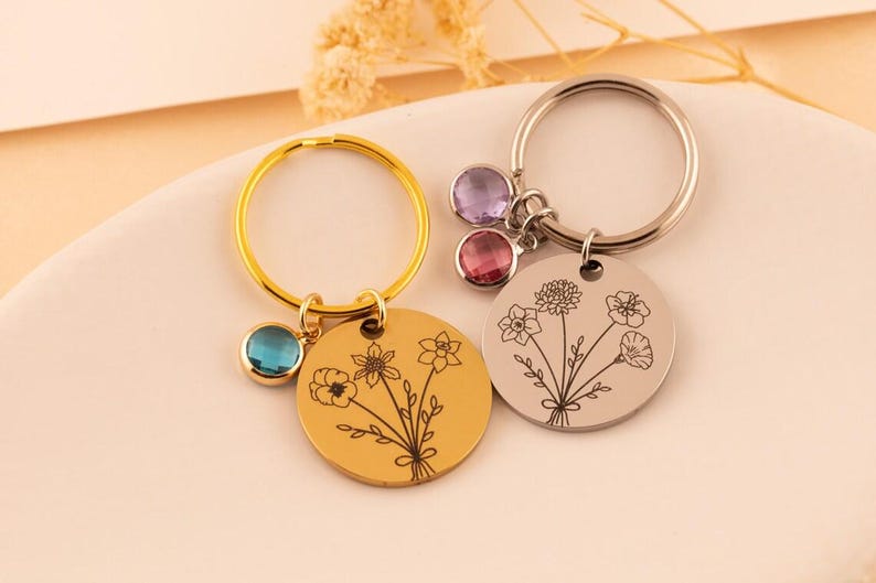 May include: Two circular metal keychains with engraved floral bouquets. One keychain is gold-toned with a blue crystal charm, and the other is silver-toned with two crystal charms in light purple and pink, featuring detailed line art of flowers.
