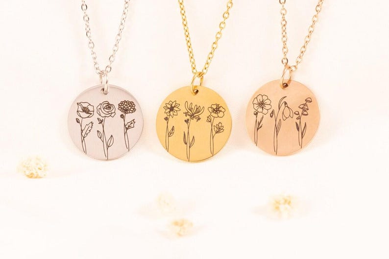 May include: Three circular pendant necklaces in silver, gold, and rose gold tones. Each round metal disc features a delicate black line art engraving of three distinct wildflowers. The pendants hang from matching thin metal link chains.