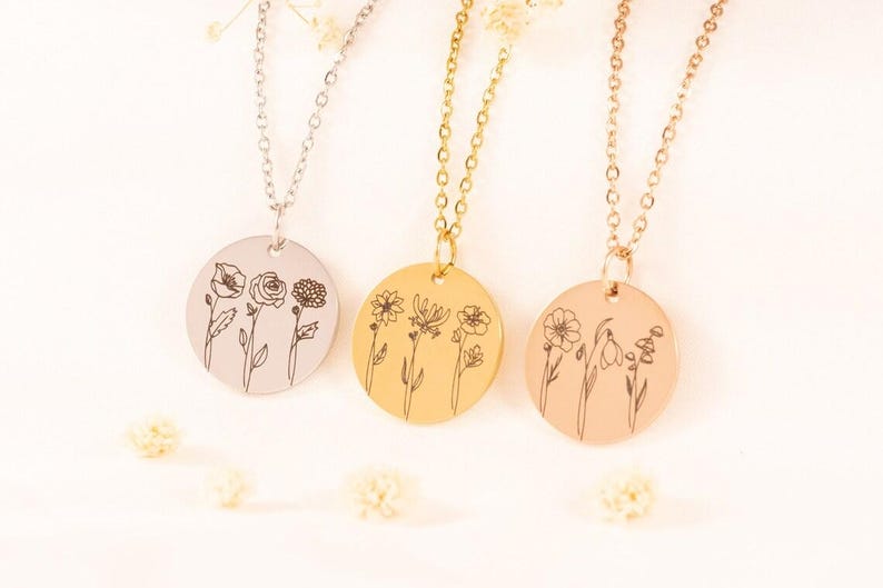May include: Three circular pendant necklaces in silver, gold, and rose gold tones. Each pendant features a delicate black line art illustration of three different wildflowers. The necklaces are suspended from matching thin metal link chains.