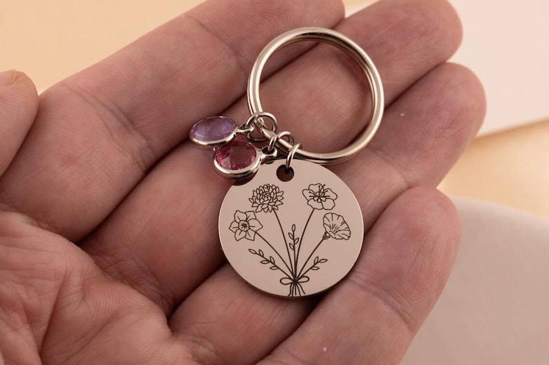 May include: A silver-toned metal keychain featuring a circular pendant engraved with a bouquet of line-art flowers. The keychain includes a split ring attachment and two small, round, faceted charms in light purple and pink hues.