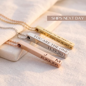 May include: Three metal bar necklaces in gold, silver, and rose gold tones. Each bar features custom engravings: one with coordinates and text, one with the phrase YOU ARE THE ONE and a date, and one with initials J and M, a heart, and a date.