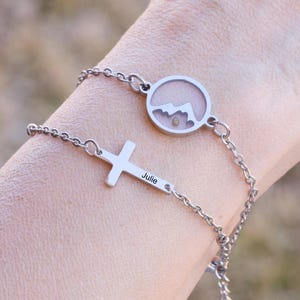 May include: Two silver bracelets on a wrist. One bracelet has a cross charm with the name "Julie" engraved on it. The other bracelet has a round charm with a mountain silhouette inside.