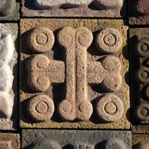 May include: A close-up of a stone relief sculpture with a cross-shaped design. The sculpture is made of brown stone and has a rough texture. The cross is surrounded by a series of circles.