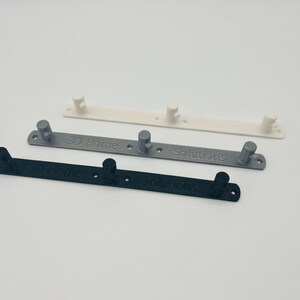 Op de afbeelding: Drie 3D-geprinte wandkapstokken in wit, grijs en zwart. Elke kapstok heeft twee of drie haken en de tekst "3D Home Solutions". De kapstokken zijn ontworpen om items op te hangen.
