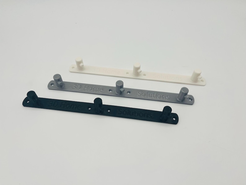 Op de afbeelding: Drie 3D-geprinte wandkapstokken in wit, grijs en zwart. Elke kapstok heeft twee of drie haken en de tekst "3D Home Solutions". De kapstokken zijn ontworpen om items op te hangen.