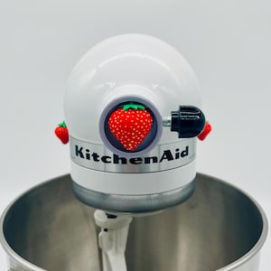 Könnte beinhalten: Ein weißer KitchenAid-Küchenmixer mit einer Edelstahlschüssel. Der Mixer hat einen großen roten Erdbeerakzent und das KitchenAid-Logo. Zwei kleine Erdbeerakzente befinden sich an der Seite.