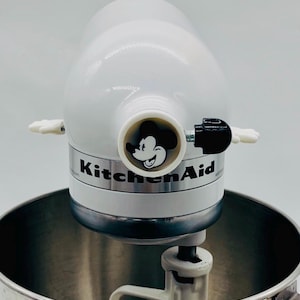 May include: A white KitchenAid stand mixer featuring a circular Mickey Mouse graphic on the front attachment hub. The mixer includes a stainless steel bowl, a white mixing paddle, and a black adjustment knob on the side of the head unit.