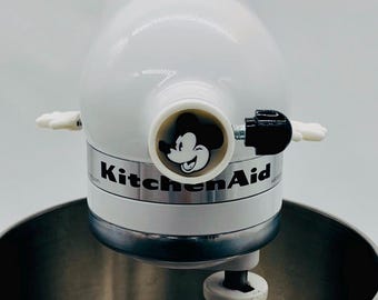 May include: A white KitchenAid stand mixer featuring a circular Mickey Mouse graphic on the front attachment hub. The mixer includes a stainless steel bowl, a white mixing paddle, and a black adjustment knob on the side of the head unit.