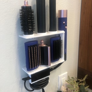 May include: White wall-mounted organizer with a blue and black hair styling tool holder. The organizer has two shelves and a bottom compartment for cords. The styling tools include a curling iron, a round brush, and a flat iron.