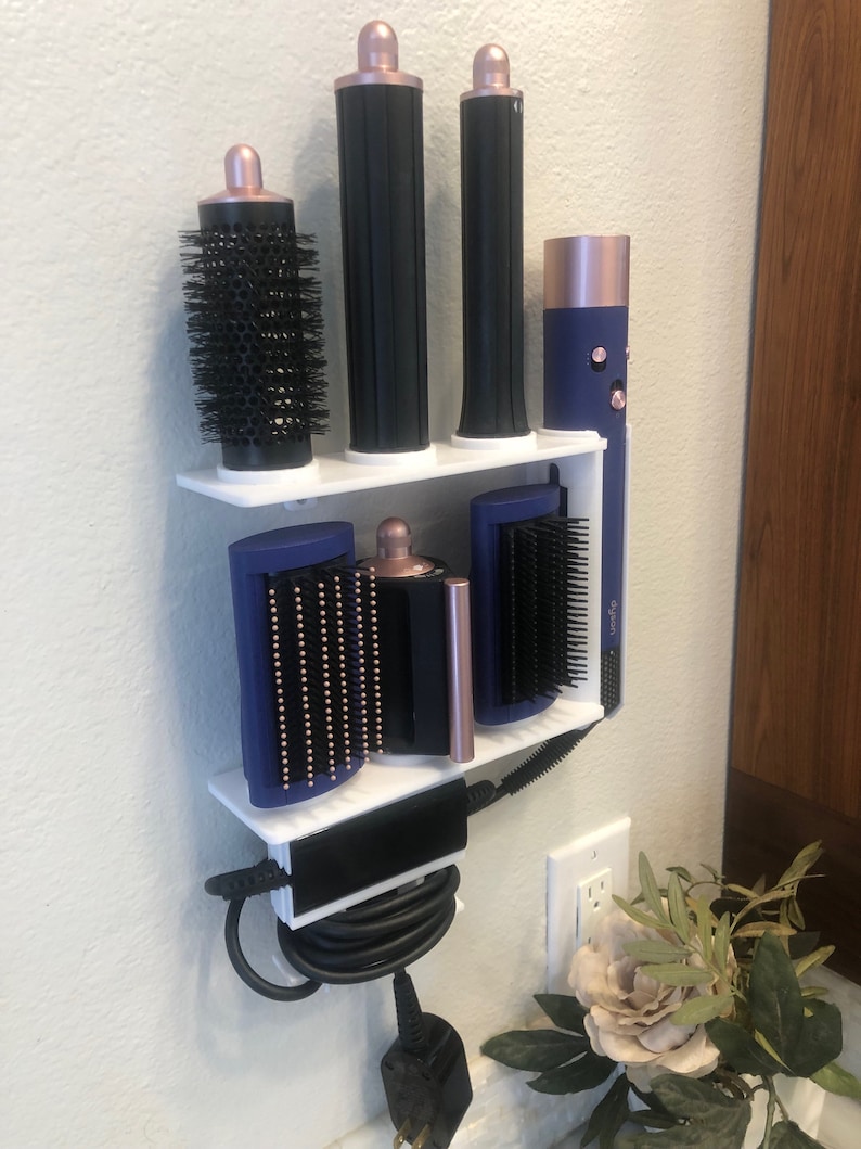 May include: White wall-mounted organizer with a blue and black hair styling tool holder. The organizer has two shelves and a bottom compartment for cords. The styling tools include a curling iron, a round brush, and a flat iron.