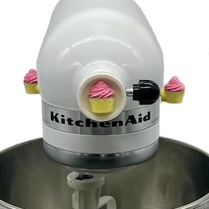 May include: A white KitchenAid stand mixer, with the brand name visible. The mixer is decorated with three cupcake-shaped ornaments, each with pink icing and a yellow base. The mixer's bowl and attachments are also visible.