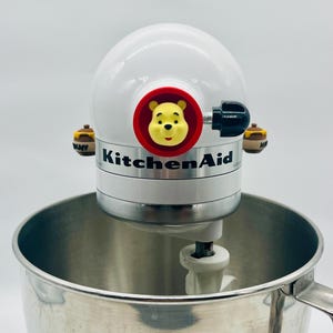 以下が含まれることがあります： 前面のハブにプーさんのキャラクターヘッドのアクセサリー、黒いコントロールノブ、側面に2つの小さなハニーポットの装飾が施された白いKitchenAidスタンドミキサー。大きなステンレス製のボウルの上に配置されています。
