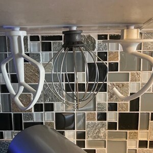 Op de afbeelding: Drie opzetstukken voor een keukenmixer: een witte platte klopper, een garde en een witte deeghaak. De opzetstukken zijn gemonteerd op een betegelde achterwand. De platte klopper heeft een uniek ontwerp met drie tanden.