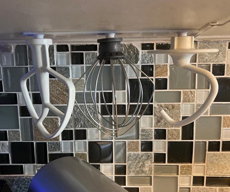 Op de afbeelding: Drie opzetstukken voor een keukenmixer: een witte platte klopper, een garde en een witte deeghaak. De opzetstukken zijn gemonteerd op een betegelde achterwand. De platte klopper heeft een uniek ontwerp met drie tanden.