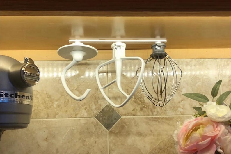 Op de afbeelding: Witte houder voor mixeropzetstukken onder de kast met een deeghaak, een platte klopper en een garde. De houder is onder een houten kast gemonteerd. Een KitchenAid-mixer is gedeeltelijk zichtbaar.