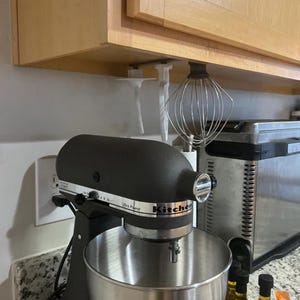 Op de afbeelding: Een zwarte keukenmixer met een roestvrijstalen kom, in een keuken. De mixer heeft de tekst "Ultra Power" en "KitchenAid". Een garde en andere hulpstukken hangen onder een kast.