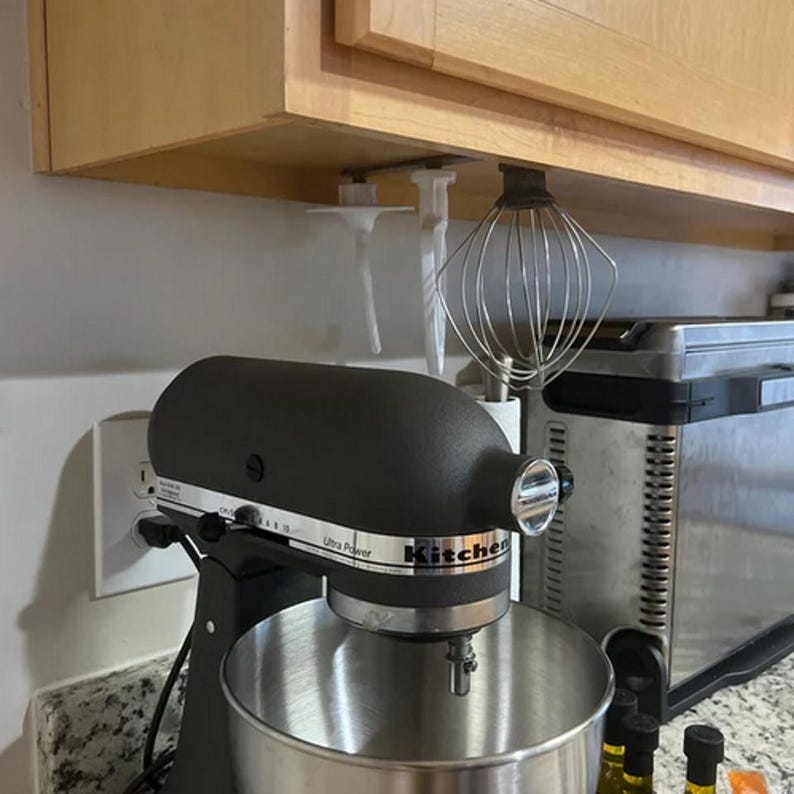Op de afbeelding: Een zwarte keukenmixer met een roestvrijstalen kom, in een keuken. De mixer heeft de tekst "Ultra Power" en "KitchenAid". Een garde en andere hulpstukken hangen onder een kast.