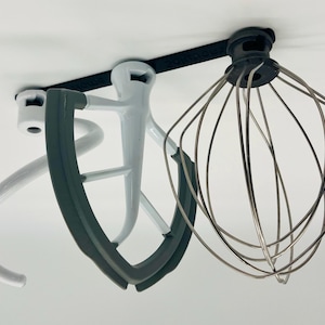 Op de afbeelding: Een verzameling opzetstukken voor een keukenmachine, waaronder een garde, een deeghaak en een platte klopper. De opzetstukken zijn in wit, grijs en zilver en zijn ontworpen voor diverse mengtaken in de keuken.