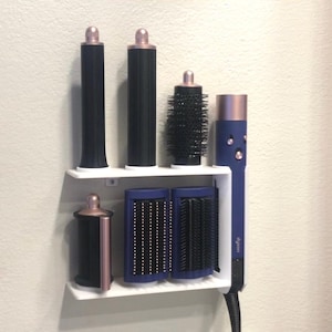 May include: White wall-mounted organizer with six hair styling tools. The organizer has two shelves and holds two black curling wands, a round brush, two blue and rose gold brushes, and a rose gold curling iron with the word "Dyson" printed on it.