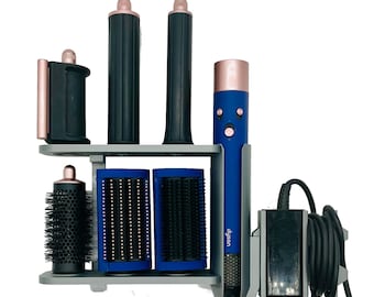 May include: A blue and rose gold Dyson hair styling tool set with multiple attachments, including curling barrels and brushes, organized on a gray wall-mounted storage rack with a black power cord and a rectangular power adapter attached to the side.