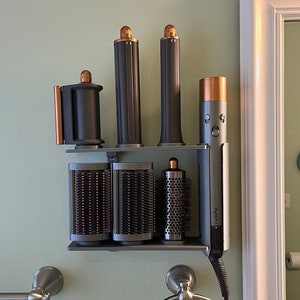 May include: A gray metal wall-mounted storage rack with four black curling irons and a Dyson hair styling tool. The curling irons have a copper-colored top and the Dyson tool has a copper-colored handle.