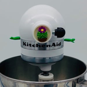 Puede incluir: Una batidora de pie KitchenAid blanca con un acento decorativo de cactus. El acento presenta un cactus verde con una flor morada y centro amarillo. La batidora tiene un cuenco de acero inoxidable y detalles verdes.