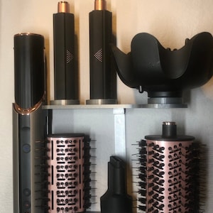 May include: A wall-mounted hair styling tool organizer with a black and rose gold hair dryer, two round brushes, a diffuser, and two cylindrical attachments.