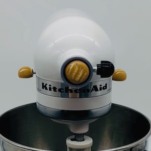 Könnte beinhalten: Ein weißer KitchenAid-Küchenmixer mit einer Edelstahlschüssel. Der Mixer hat ein weißes Gehäuse, ein silbernes Band und das KitchenAid-Logo. Er verfügt über Holzknöpfe und einen schwarzen Steuerhebel. Die Schüssel ist aus Edelstahl.