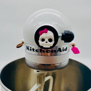 May include: A white KitchenAid stand mixer featuring a decorative skull emblem with a pink bow on the front. The mixer includes a silver metal band with the KitchenAid logo, a white mixing attachment, and a large stainless steel mixing bowl.