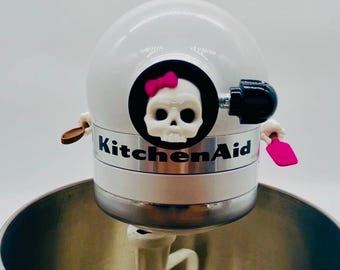 May include: A white KitchenAid stand mixer featuring a decorative skull emblem with a pink bow on the front. The mixer includes a silver metal band with the KitchenAid logo, a white mixing attachment, and a large stainless steel mixing bowl.